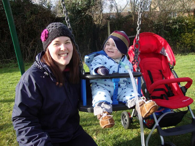 Mum smiling to the camera beside her baby on a swing attached to a breathing tube