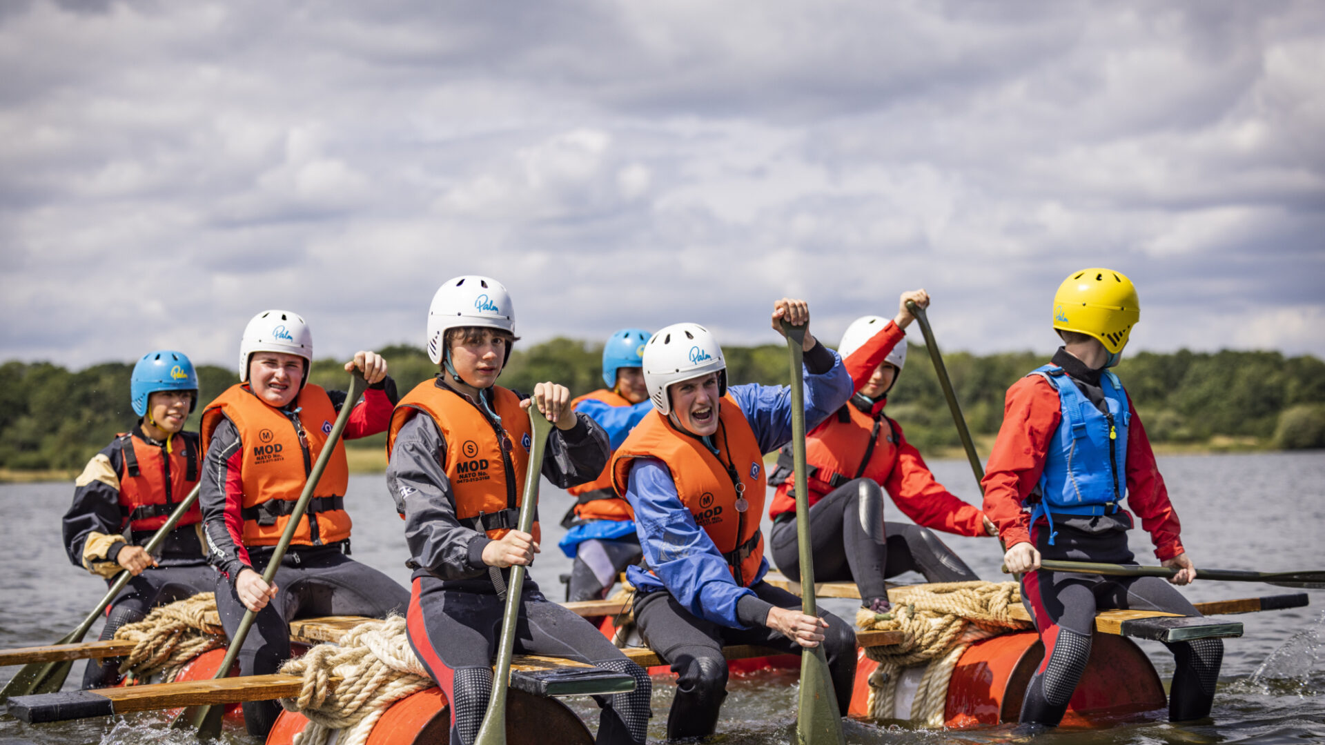 seven teenagers wearing orange life jackets paddling on a raft across a lake, putting in lots of effort.