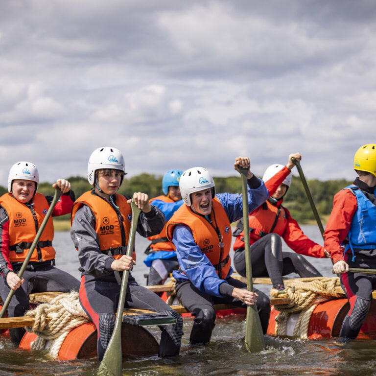 seven teenagers wearing orange life jackets paddling on a raft across a lake, putting in lots of effort.