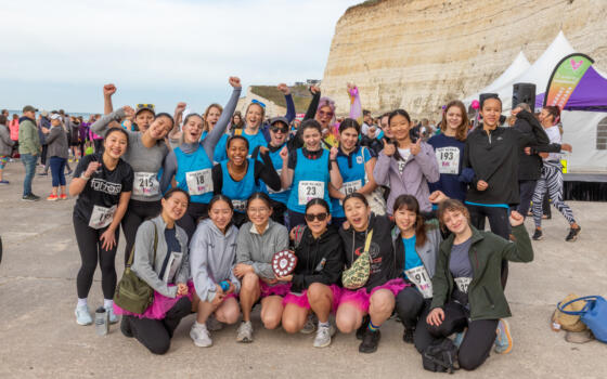 A colour photo of 22 girls and women grouped together on the Undercliff path, wearing running clothes, race numbers and some in pink tutus, with fists in the air smiling celebrating completing their run