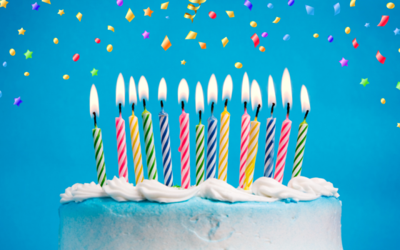 A white birthday cake with lit multicoloured candles on a blue background