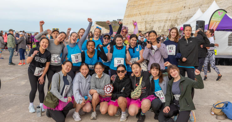 A colour photo of 22 girls and women grouped together on the Undercliff path, wearing running clothes, race numbers and some in pink tutus, with fists in the air smiling celebrating completing their run