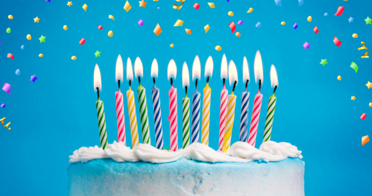 A white birthday cake with lit multicoloured candles on a blue background