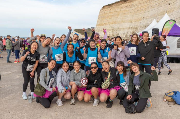 A colour photo of 22 girls and women grouped together on the Undercliff path, wearing running clothes, race numbers and some in pink tutus, with fists in the air smiling celebrating completing their run