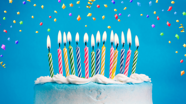 A white birthday cake with lit multicoloured candles on a blue background