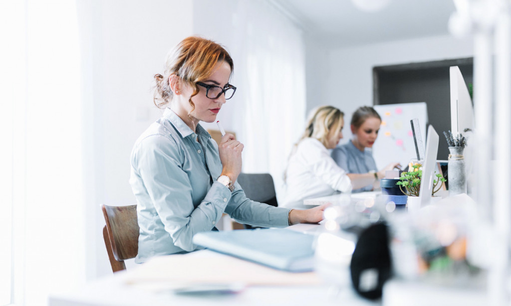 Focused woman in glasses working at desk with laptop, while two colleagues collaborate in the background | Amodaia
