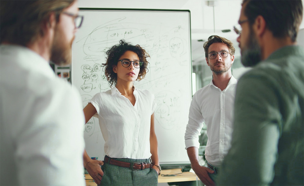 Confident businesswoman in white shirt and glasses leading a serious team discussion in front of a whiteboard filled with handwritten notes and diagrams | Amodaia Confident businesswoman in white shirt and glasses leading a serious team discussion in front of a whiteboard filled with handwritten notes and diagrams | Amodaia