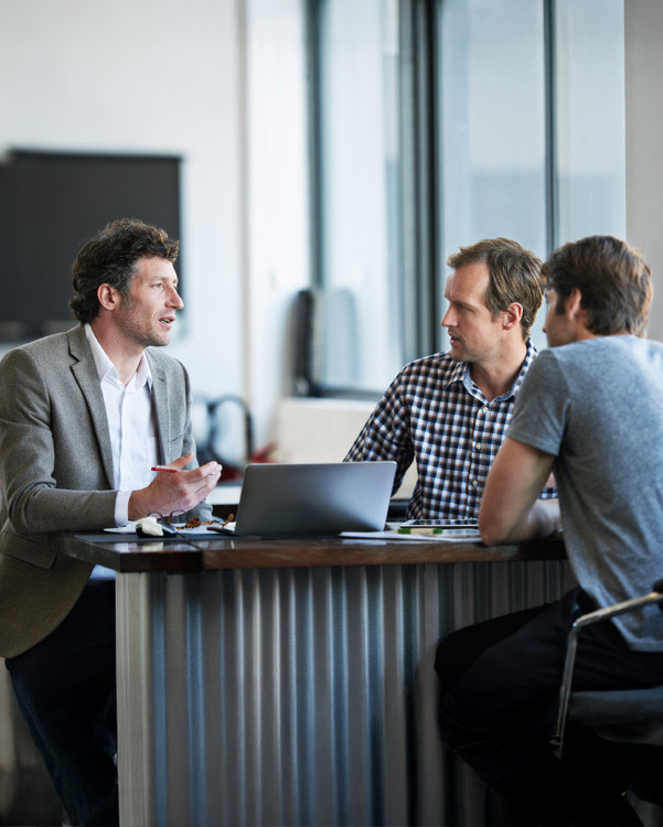 Three colleagues in conversation around a laptop at a high table in a modern office | Amodaia