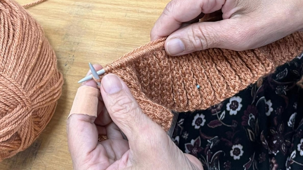 Close-up of hands knitting with orange yarn and needles, ball of wool resting on a wooden table | Catherine Beckett Centre