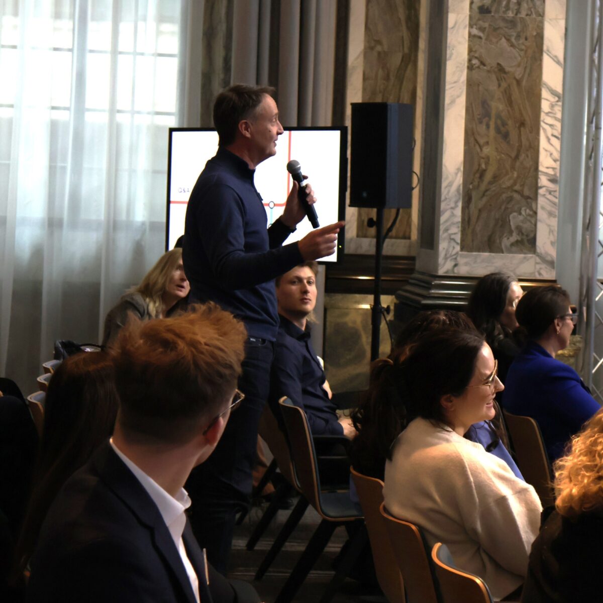 A volunteer stands among seated attendees during a panel event, holding a microphone and speaking while the audience listens, in a bright conference venue with tall windows, marble columns, and presentation screens in the background.