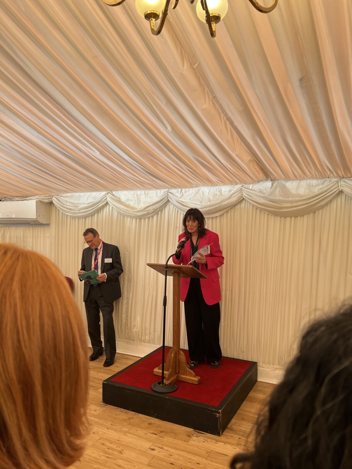 Woman in a red jacket standing behind a board making a speech.
