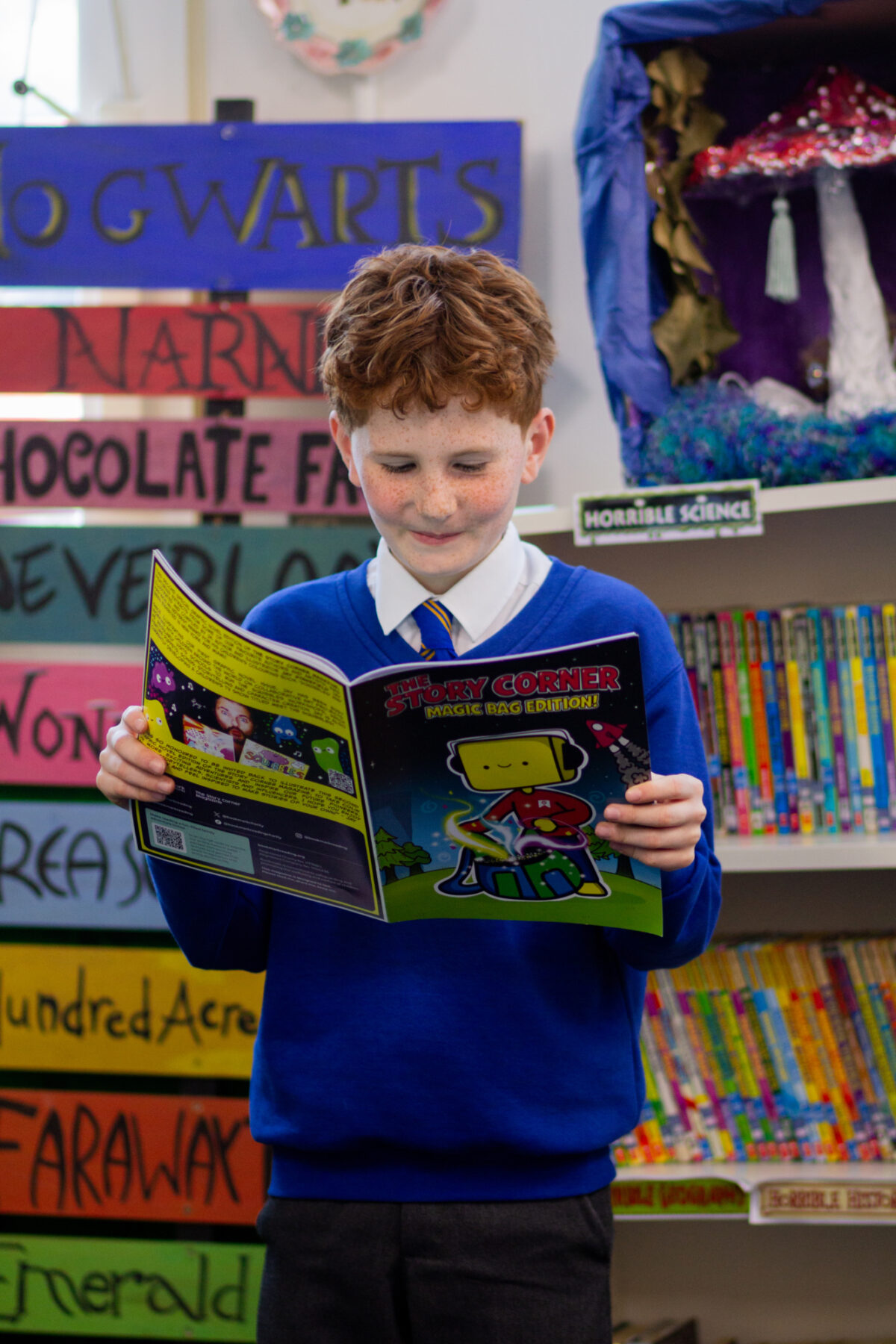 Child from Morden Primary School holding Bookmark's The Story Corner magazine. He's surrounded by shelves of colourful children's books.