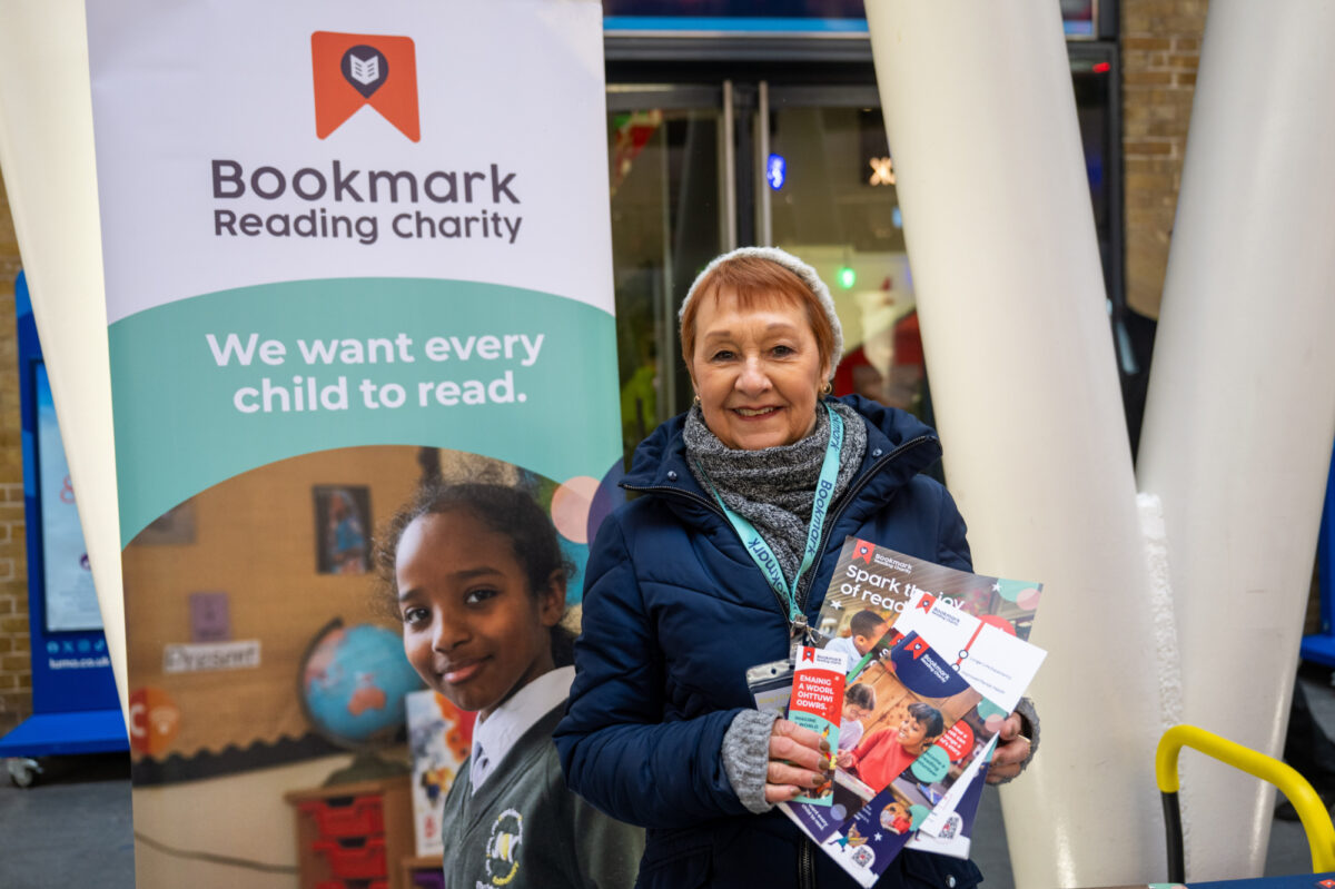 A volunteer stands at a Bookmark Reading Charity stall, smiling and holding a children’s book, with charity signage behind them that reads “We want every child to read.”  Mind the Gap volunteer activation at King’s Cross London