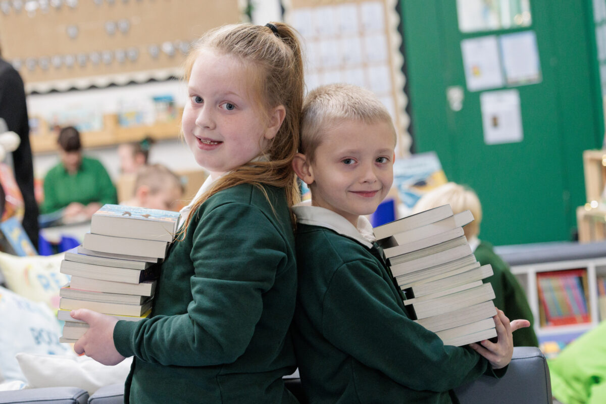 Two girls standing back to back smiling at the camera whilst holding a pile of books from the Roots to reading programme