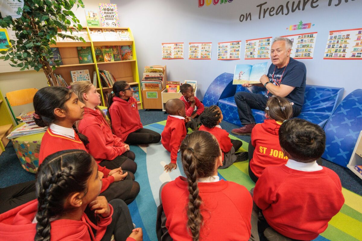 Children's author is reading to a small group of children who are sat on the floor. Author is showing them a page in the book.