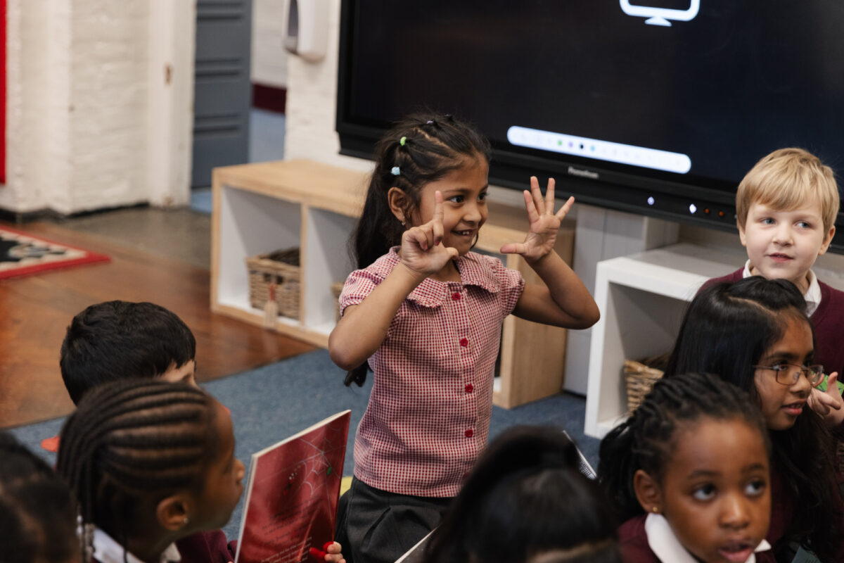 A young child stands and gestures with both hands while participating in a classroom activity, surrounded by other children seated on the floor.