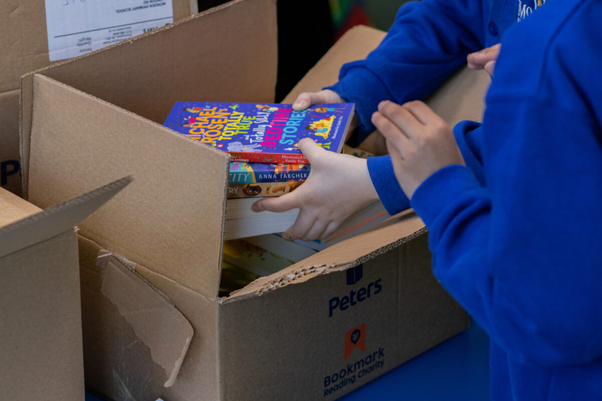 Children in blue school uniforms unpacking colourful storybooks from a large Peters delivery box during a Roots to Reading session at Morden Primary School. A pair of hands is lifting a stack of books from the box while another child reaches in to help organise the newly delivered reading materials