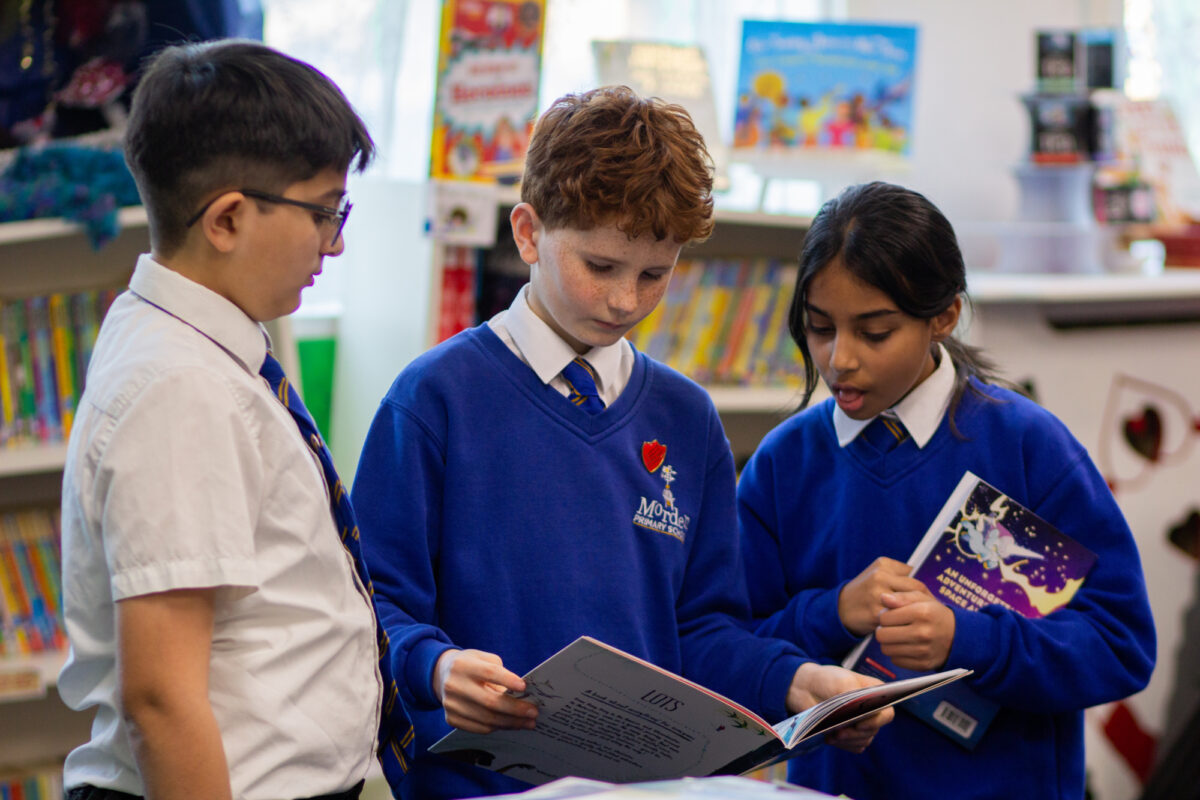Three children at Morden Primary School standing together in the library as part of the Roots to Reading programme. The children are holding open books, reading and discussing the pages with one another while surrounded by shelves of colourful children’s books.