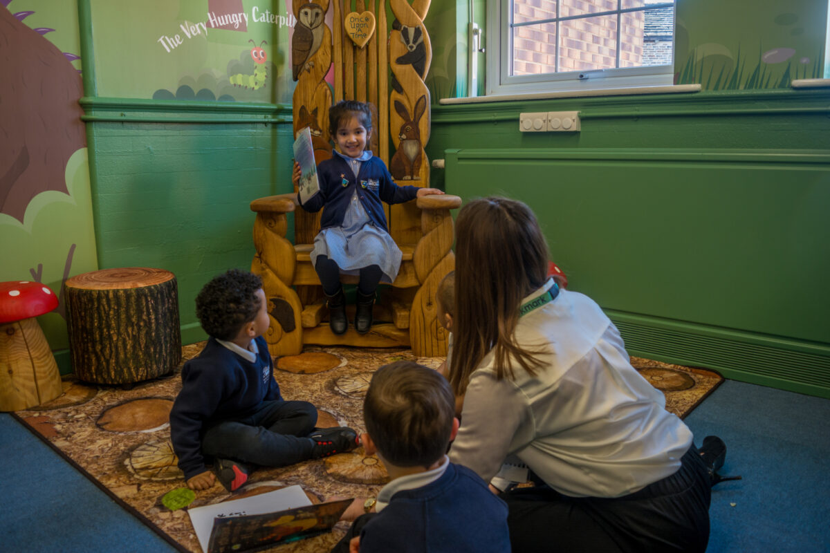 Girl sat in wooden chair holding a book, with two children and teacher sat on a mat in front of her