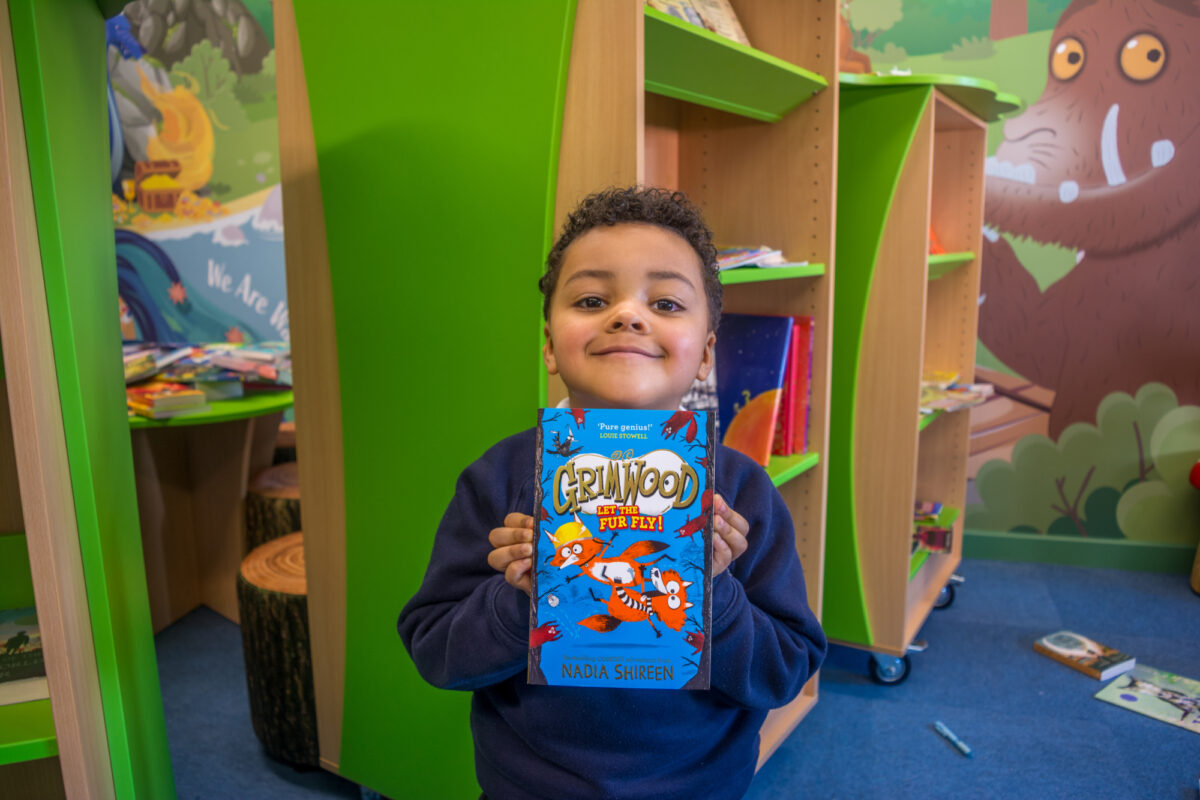 Boy in blue school uniform, smiling and holding up a book.