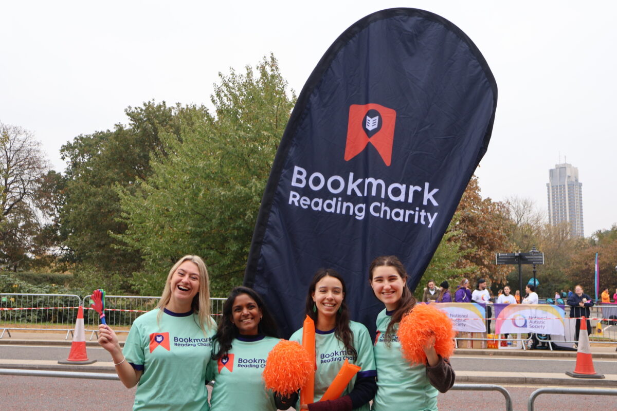 4 women smiling at the camera, they're wearing turquoise Bookmark t-shirts and cheering with pom poms and clappers in front of a Bookmark flag
