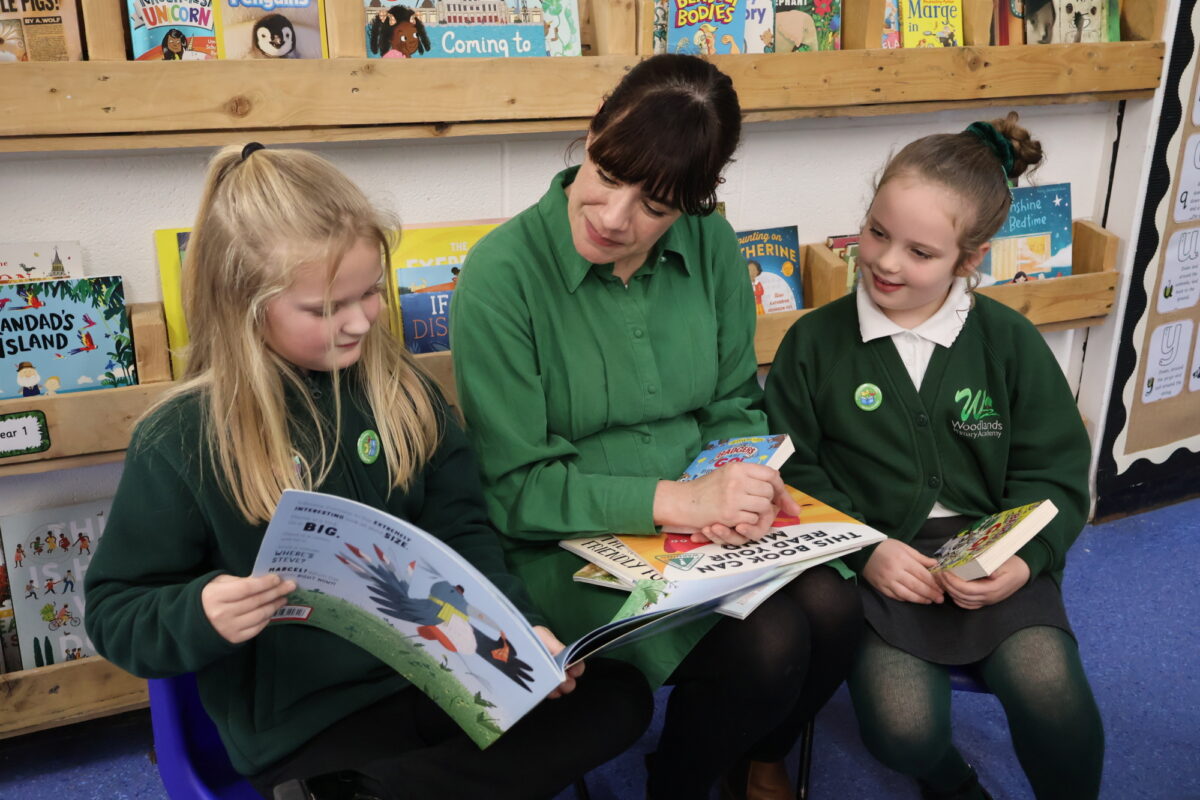 Author reading with two pupils, she has a book on her lap and one of the girls is pointing to a page.