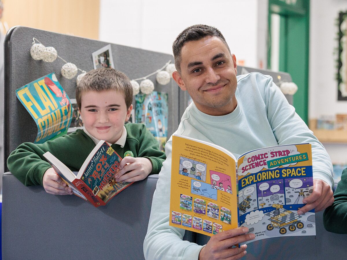 Two people sitting on a gray bench reading books, one titled “Exploring Space” and the other “The Brilliant World of Tom Gates,” with posters and string lights in the background.