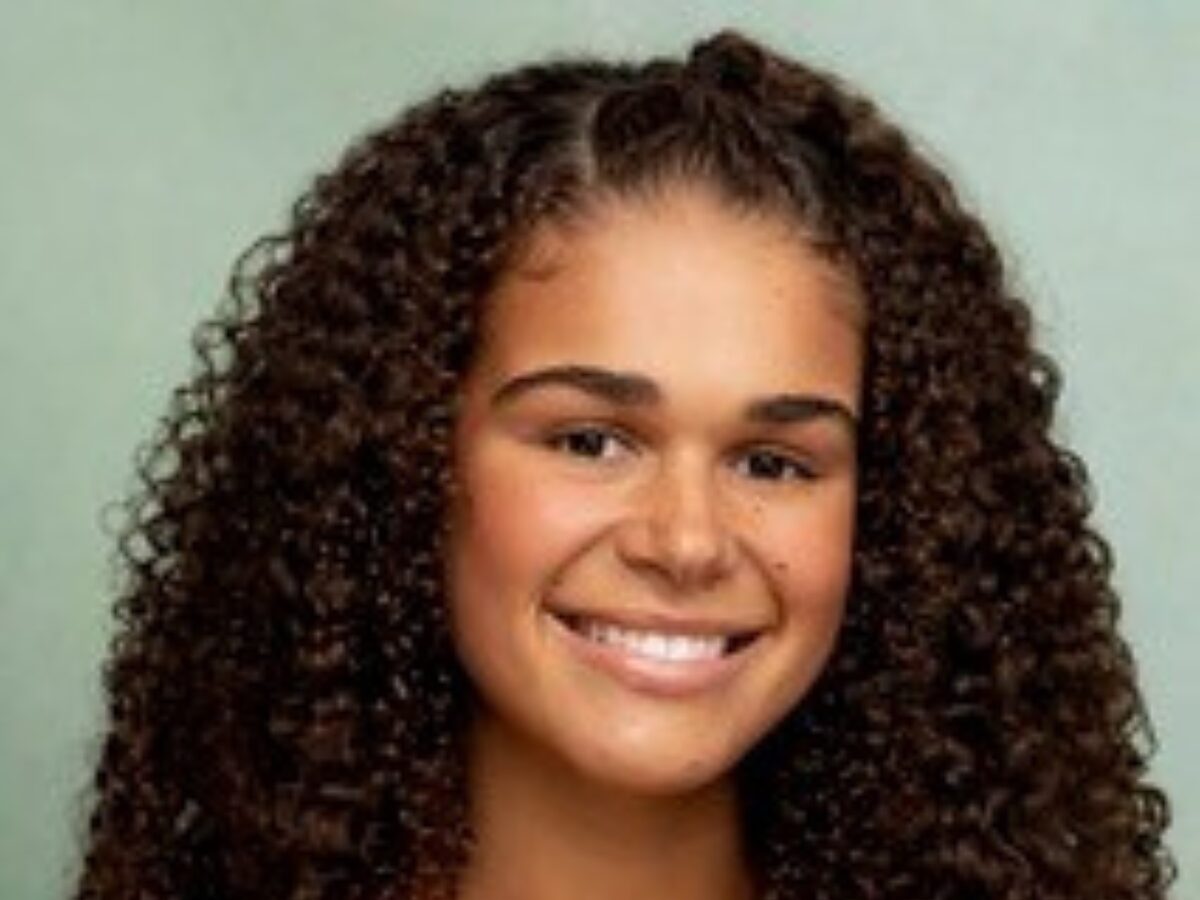 Person with long, dark curly hair wearing an orange collared shirt, positioned against a plain light green background.