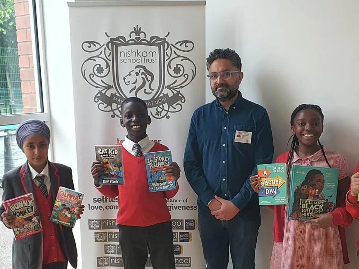 Four people standing indoors in front of a Nishkam School Trust banner, with three holding colorful children’s books including “Bunny vs Monkey,” “Olivia,” “Cat Kid Comic Club,” “26-Storey Treehouse,” “A Better Day,” and “Brilliant Black British History.”