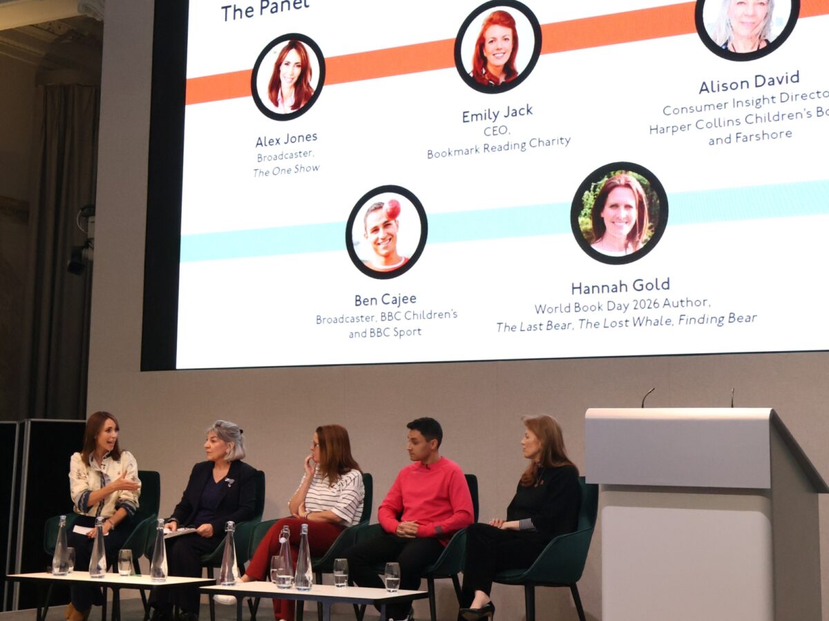 Five speakers sit on stage for a panel discussion, with a large presentation slide behind them displaying the event’s panel members