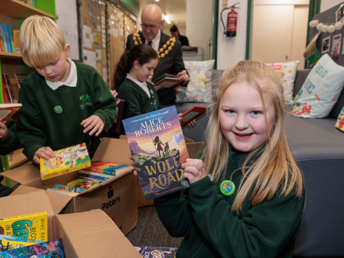 A girl in a green jumper holds up a book, while other children open boxes of books in the background