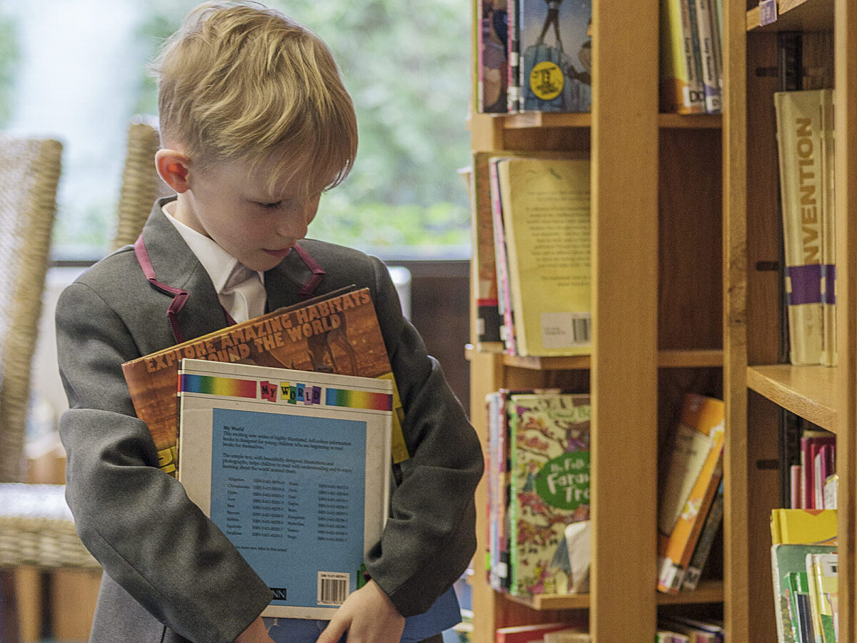 Boy with blonde hair in school uniform holding a variety of books, looking at the library shelves