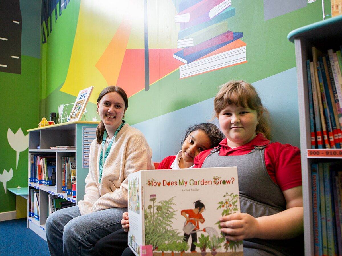 Two children sat with a volunteer, they're wearing red school uniform and are holding up a book.