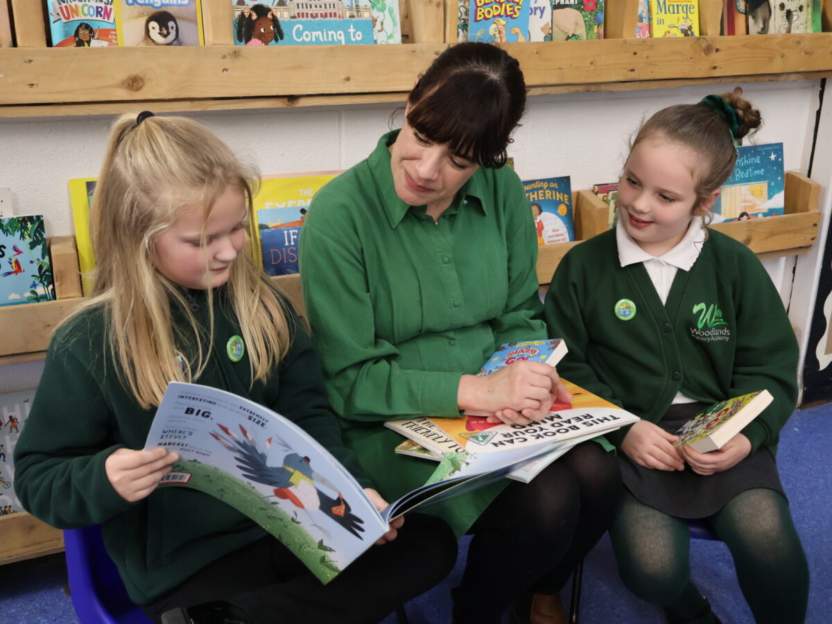 Author reading with two pupils, she has a book on her lap and one of the girls is pointing to a page.
