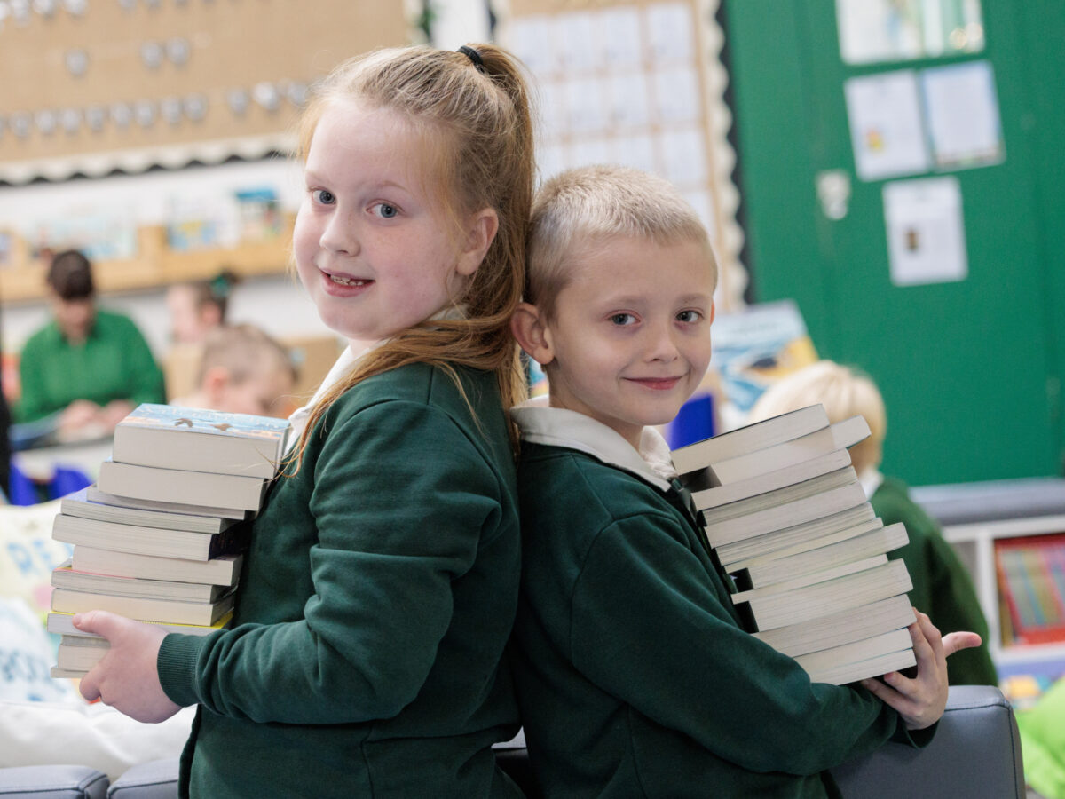 Two girls standing back to back smiling at the camera whilst holding a pile of books from the Roots to reading programme