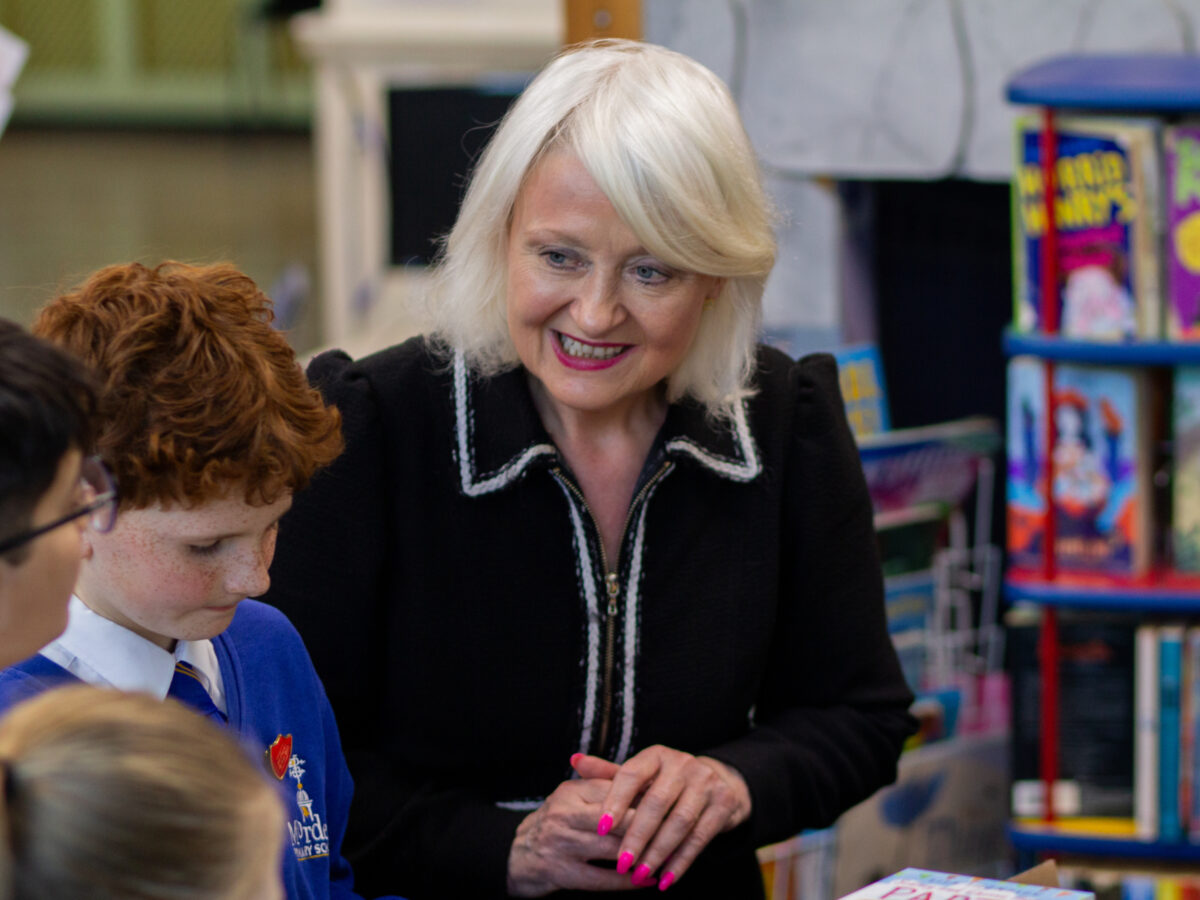 During a Roots to Reading visit at Morden Primary School, a group of children gather around Merton's MP, Siobhan Mcdonagh who is standing at a table covered with books.