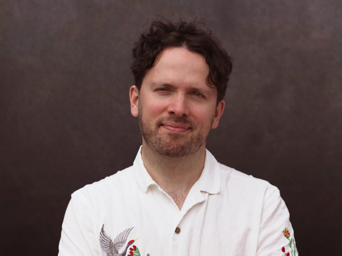 Man with curly brown hair with his arms crossed, wearing a white shirt with flowers.
