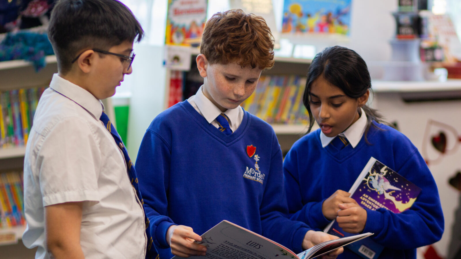 Three children at Morden Primary School standing together in the library as part of the Roots to Reading programme. The children are holding open books, reading and discussing the pages with one another while surrounded by shelves of colourful children’s books.