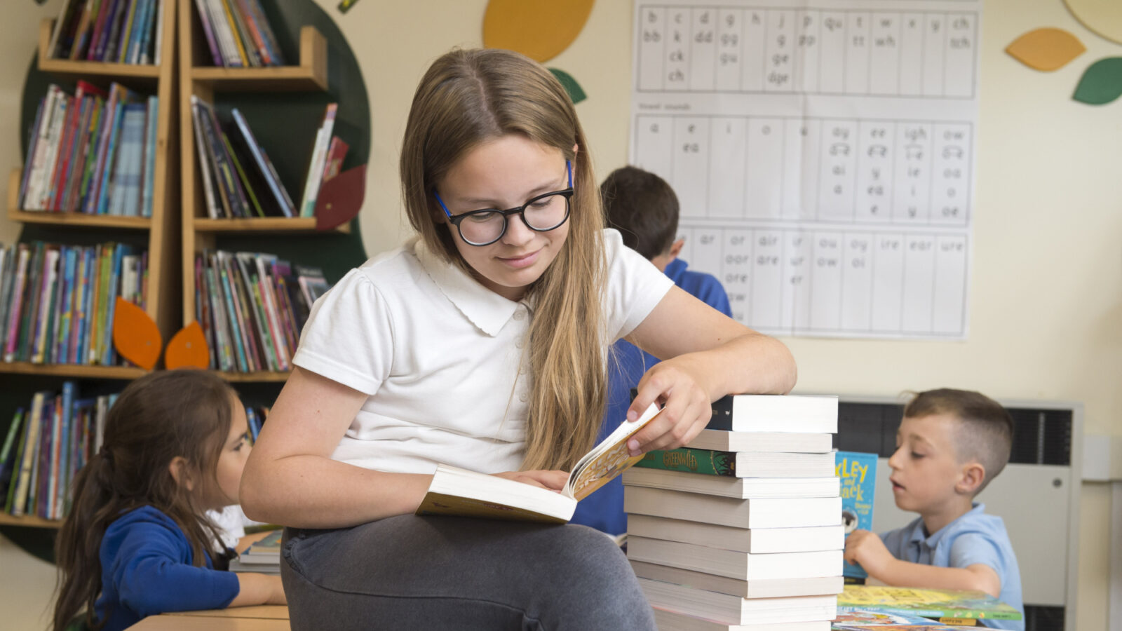 A girl in a white top reads a book, leaning her arm on a stack of books