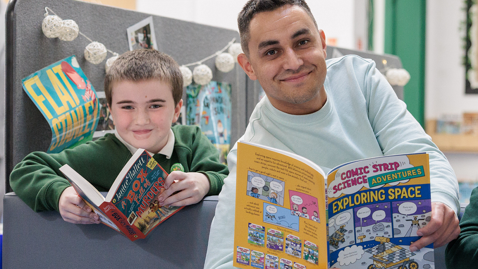 Two people sitting on a gray bench reading books, one titled “Exploring Space” and the other “The Brilliant World of Tom Gates,” with posters and string lights in the background.