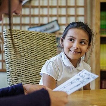 Child in a white school uniform smiling at a Bookmark reading volunteer holding her drawing during a one-to-one reading support session.