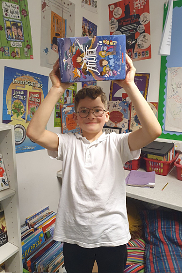 Boy holding up graphic novel he is smiling at the camera and is wearing white school uniform