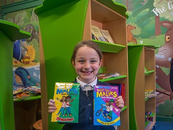 A young girl in a school library smiling holding two books in front of her