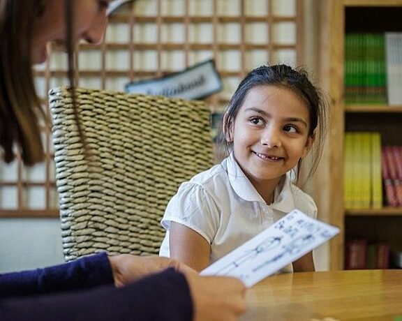Child in a white school uniform smiling at a Bookmark reading volunteer holding her drawing during a one-to-one reading support session.