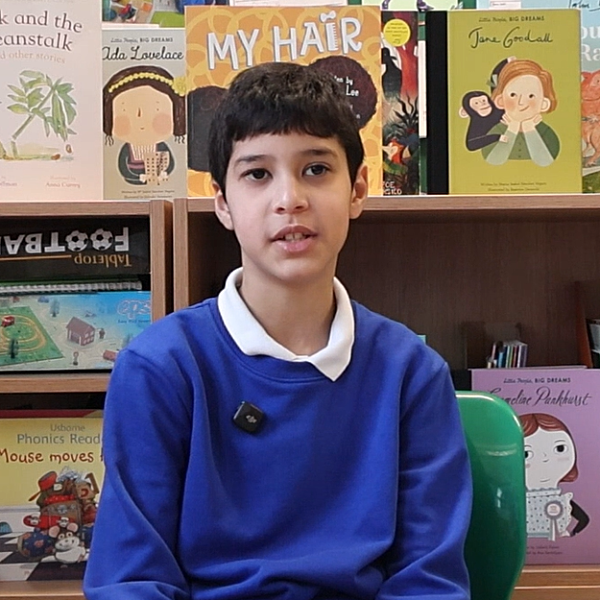 Boy in school uniform sits talking to the camera with a library of books behind.