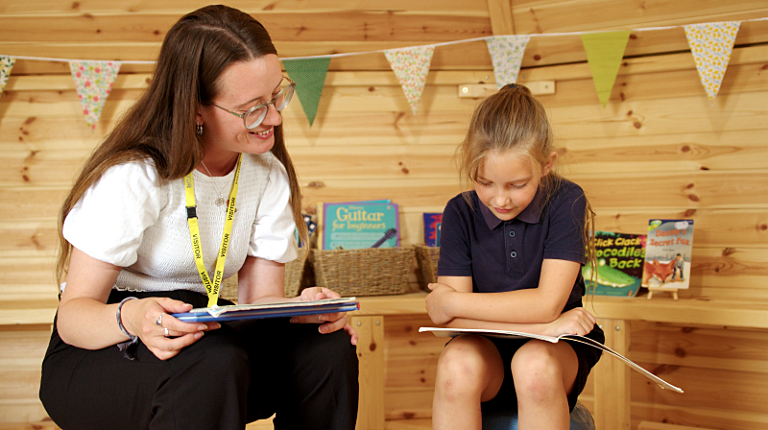 A reading volunteer supporting a child during a one‑to‑one reading session in a cosy, book‑filled space.