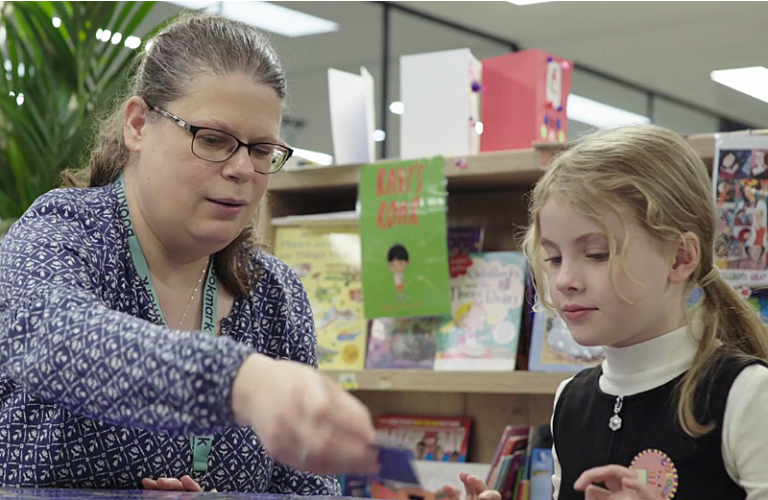 Claire has long grey hair tied back in a ponytail, she is wearing a purple top and a bookmark lanyard. she is playing literacy cards with her reader