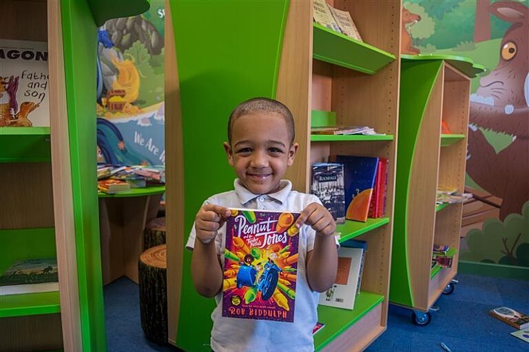 A young boy in a school library smiling holding a book in front of him