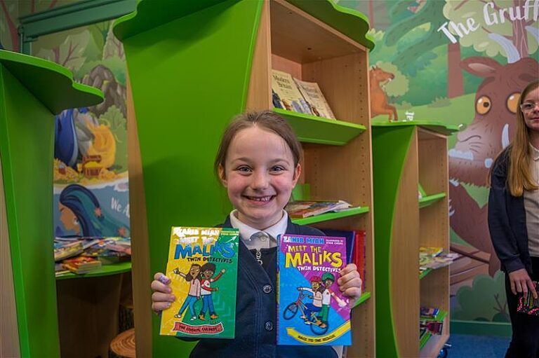 A young girl in a school library smiling holding two books in front of her