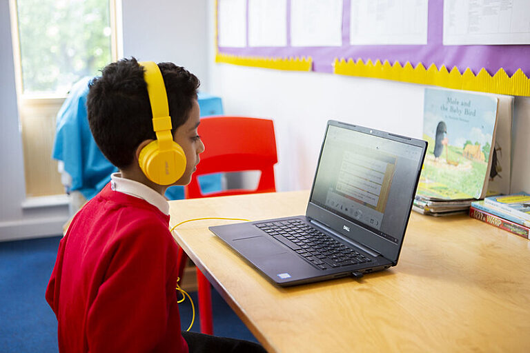 A child wearing headphones taking part in an online reading session on a laptop in a classroom setting.